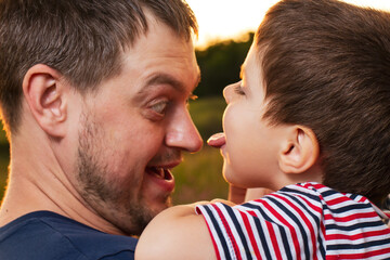 The son licks his father's tongue in the nose. Games of child and adult, love, Father's Day. Dad and baby play on the back of the field.