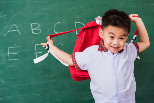 Back To School. Happy Asian Funny Cute Little Child Boy Kindergarten Preschool In Student Uniform Wearing School Bag Stand Smiling On Green School Blackboard, First Time To School Education Concept