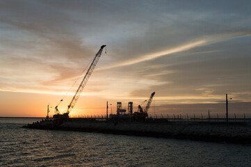 Fototapeta na wymiar オレンジ色の夕暮れの空をバックに作業している海岸沿いのシルエットのクレーン