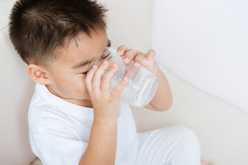 Close up of happy Asian little cute child boy hand holding milk glass he drinking white milk during sitting on the sofa at home after lunch. Daily life health care Medicine food