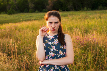 A girl in a summer yellow field eats candy on a stick and emotionally frowns. Evening in the field and a woman.