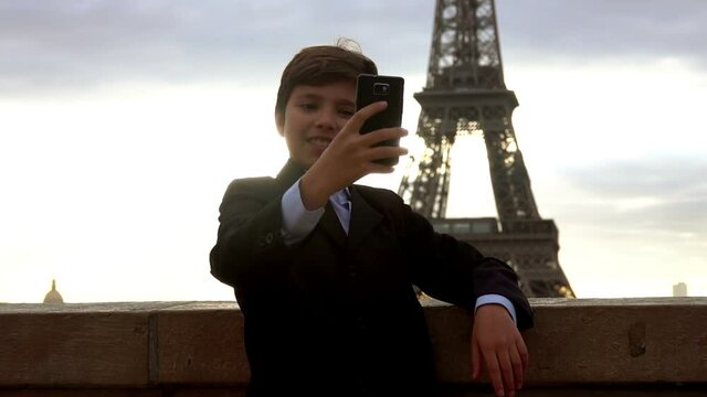 Handsome schoolboy in a suit is making a selfie on the phone on the background of the Eiffel tower at the dawn, Paris, France