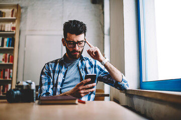 Young confused hipster guy in spectacles reading text message from work