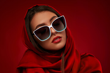 Portrait of chic brunette woman with luxurious make-up in white sunglasses and red headscarf on red...