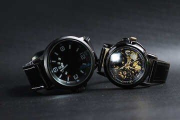 a black automatic self winding wristwatch with transparent sekeleton dial design and a black battery operated watch on black leather background