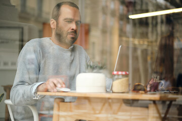 Fototapeta na wymiar Thoughtful casual businessman intently working on laptop in cafe. Window view