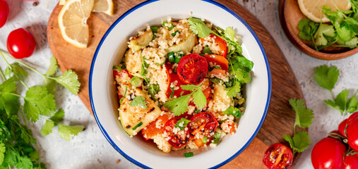 Salad of couscous and grilled vegetables in the bowl, cilantro, lemon and tomatoes on a gray concrete background top view. Vegan food. Arab or Middle Eastern cuisine.