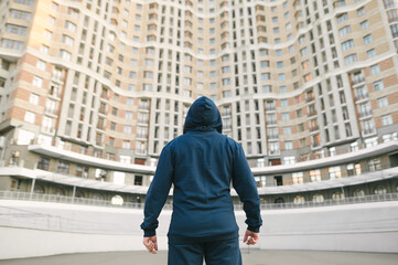 Back of an athletic man in a hood and sportswear stands after training against the background of architecture and looks up. Strong muscular man stands with his back to the building.