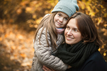 Girl hugs mom, they are smiling. In the background is the soft focus of golden orange autumn. They are wearing a jacket, hat and coat. Close-up portrait