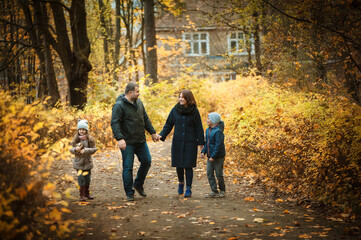 Fototapeta premium Sweet family walks along the path in the autumn park. Husband and wife hold hands, son and daughter walk together, they smile and talk. people are warmly dressed, they are in a good mood. Soft focus