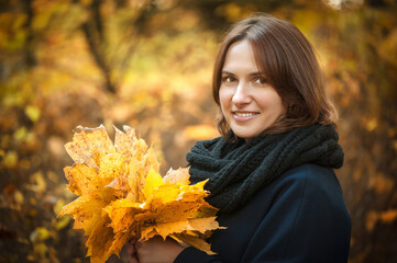 Young beautiful girl stands with a bouquet of maple orange leaves in the autumn park. They are looking at the camera and smiling. Dressed in a dark coat and black scarf. Close-up, soft focus.