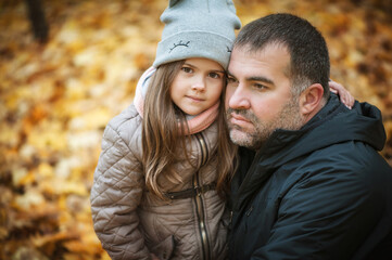 A little girl hugs her dad, they are in the autumn park, in the background soft focus autumn leaves. A man in a jacket, a girl in a gray hat.