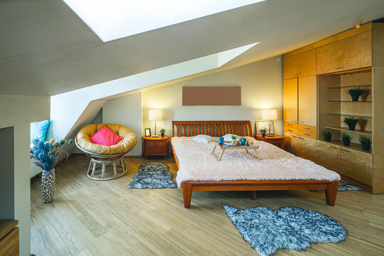 Modern interior of bedroom in apartment. Wooden furniture. Front view of bed. Sloping ceiling with window.