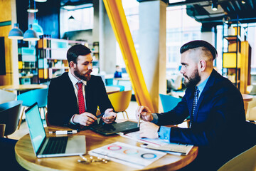 Two business partners dressed in stylish formal wear communicating with each other and discussing new productive strategy of developing common corporate project sitting at meeting table in office