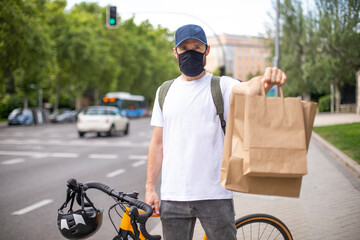 Repartidor de comida a domicilio con mascarilla en bicicleta amarilla por la ciudad con la mochila...