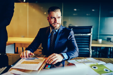 Confident male entrepreneur in formal wear sitting at his desktop with papers and documentation,...