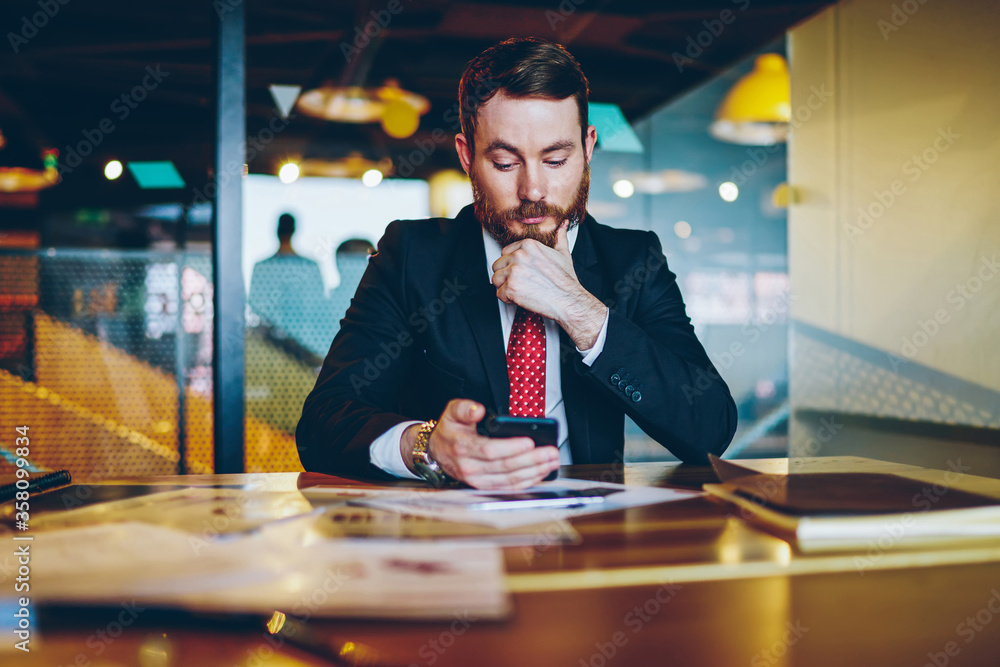 Poster pensive male entrepreneur sending text message via smartphone during working process at desktop, con