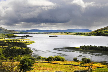 Vue panoramique sur un lac et ses &icirc;lots de terre, avec une ruine de tour m&eacute;di&eacute;vale en son centre. Ciel charg&eacute; et nuageux, collines, verdure et rochers.