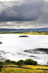 Vue panoramique sur un lac et ses îlots de terre, avec une ruine de tour médiévale en son centre. Ciel chargé et nuageux, collines, verdure et rochers.