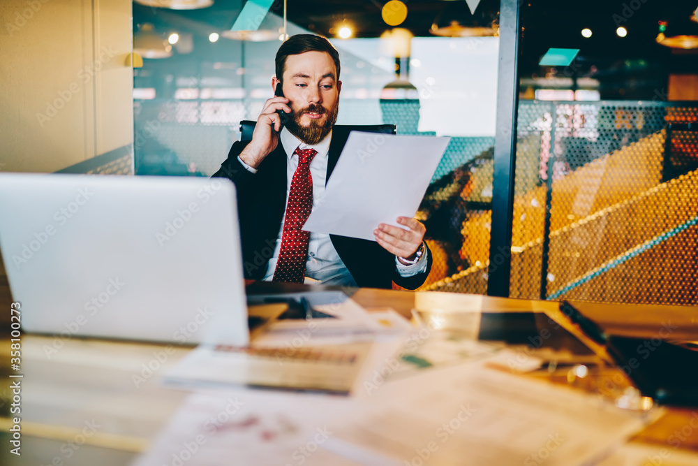 Poster bearded experienced entrepreneur in formal wear reading financial reports and calling to manager on 