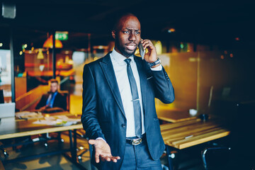 Dissatisfied African American businessman dressed in formal wear solving problem during phone conversation on smartphone.Angry dark skinned entrepreneur calling on cellular in modern office interior