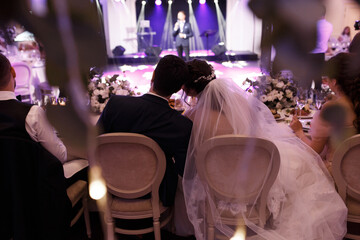 the couple is sitting at the wedding table embracing. An entertainer appears on the stage. View from the back. 