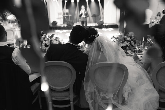the couple is sitting at the wedding table embracing. An entertainer appears on the stage. View from the back. black white image