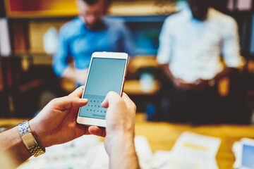 Cropped view of male hands with luxury watches holding modern smartphone device with business people on background. Selective focus on mock up screen of mobile phone,  online banking application