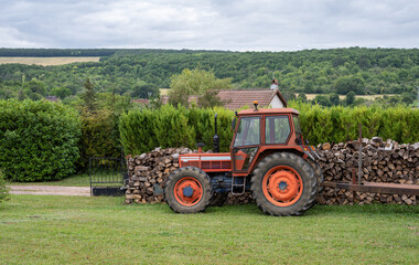 Obraz premium Red tractor parked in a garden in front a stack of cut wood. Countryside landscape.