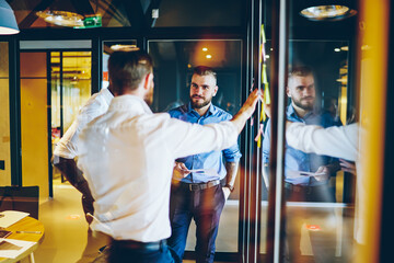 Group of entrepreneurs dressed in formal wear cooperating on success of business during meeting in modern office.Creative employees discussing working strategy standing near glass wall sharing ideas