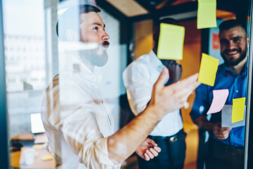 Obraz premium Cheerful multiracial colleagues discussing information on stickers on window