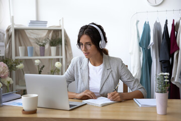 Focused Caucasian female designer in headset watch webinar on laptop making notes in notebook,...