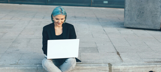 Lovely geek with blue hair working at the computer outside in the street sitting on the ground with...