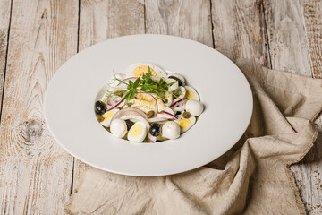 Mediterranean salad with squid rings, red onions, mozzarella, olives, quail eggs and capers on a white plate.