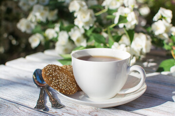 White porcelain cup with tea and two oatmeal cookies with sesame seeds on a wooden table against the background of blooming jasmine.