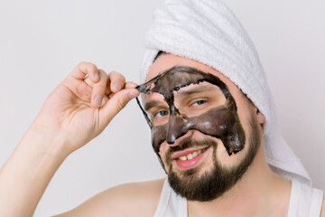 Handsome smiling bearded guy is removing purifying black mask from his face over white background. Close up portrait of man during spa treatment