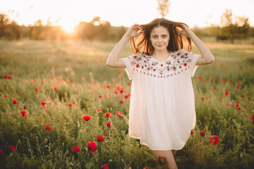 Naklejka na ściany i meble Young happy smile caucasian girl staying at a green red poppy field in the morning. The background of the sun shines. Concept of freedom. Summer. Beautiful young woman walking on field