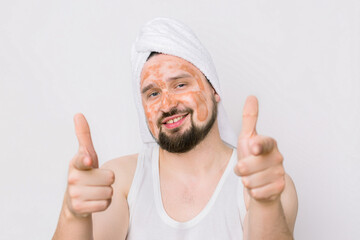 Portrait of a handsome smiling relaxed young man with a facial mud clay mask, pointing his fingers to camera. Face and body skin care treatment for men. Isolated on white