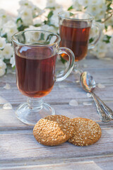 Two glass glasses with tea and two oatmeal cookies with sesame seeds on a wooden table against the background of blooming jasmine.