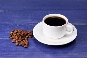 A cup of coffee and coffee beans on a blue wooden background. Heap of coffee beans. White coffee cup and saucer on a blue table.Copy space. Place for text.