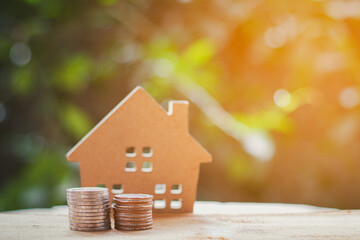 A pile of coins and wooden toy house on a natural green blur background in the morning.