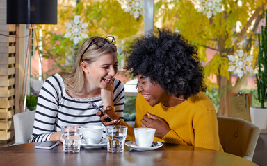 Two young girl friends enjoying coffee together in a coffee shop, sitting at a table, chatting and laughing out loud while tracking social media on a smartphone. Diversity and multi ethnic concept. 