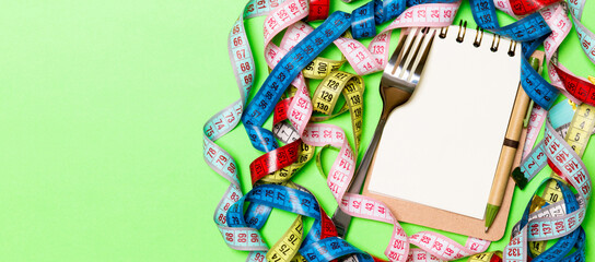 Composition of colorful measuring tapes, fork, pen and notepad on green background. Top view of diet control and empty space for your design