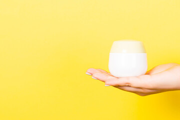 Close up of female hand holding cosmetics jar at yellow background with copy space