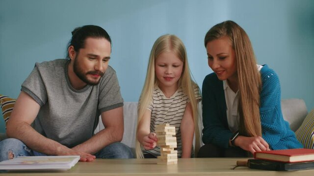 Small blonde girl plays jenga with mom and dad at home, trying to do her best, but fails, enjoys playing the game, positive emotions, Slow motion.