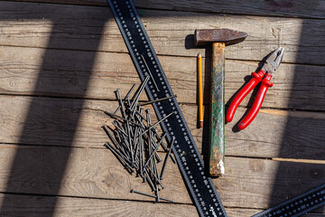 Old construction tools, nails, a hammer, a pencil, a ruler, and pliers lay on a plank wooden floor. A game of shadow and light. Repair, construction, carpentry workshop. Close-up, group of objects.