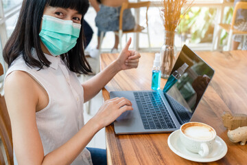 Asian woman in medical mask showing thumb up working on computer laptop with alcohol spray bottle stay at cafe. Social distance concept