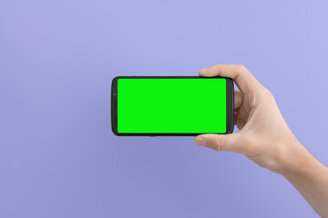Smartphone with a green screen in right hand on an lilac background, horizontal position.