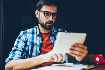 Pensive bearded young man reading news on website via 4G internet connected to digital tablet.Concentrated male blogger in eyeglasses for vision correction downloading publication on touch pad device