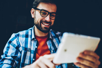 Cheerful bearded hipster guy in eyeglasses watching tutorial on digital tablet connected to 4G internet.Positive male blogger reading funny comments under publication on modern touch pad device